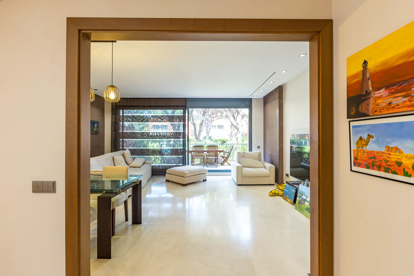 View through a doorway into a bright living room with a sofa, chair, and glass table. Artwork hangs on the wall.