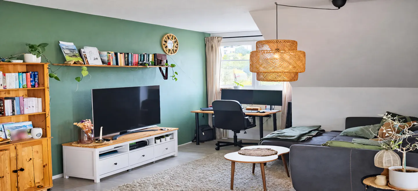 Living room with green walls, a TV, a desk with two monitors, and a gray sofa. A woven pendant light hangs from the ceiling.