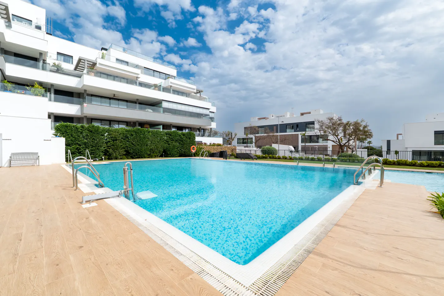 Outdoor pool with blue water and light wood deck. Modern white buildings and green hedges in the background under a cloudy sky.