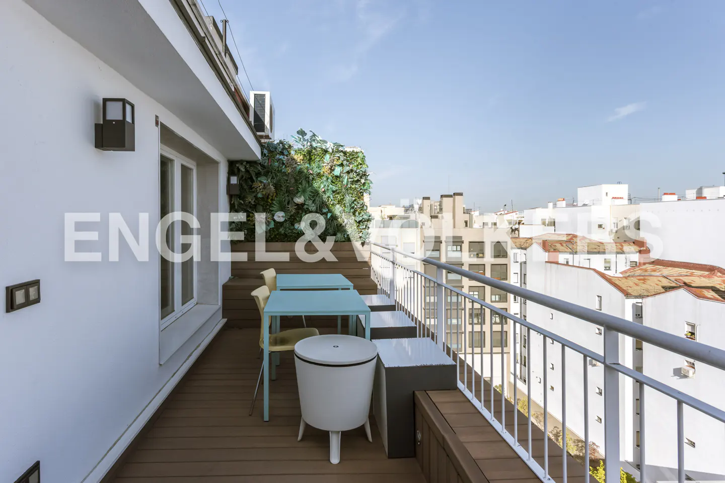 Modern rooftop terrace with wooden decking, a light blue table, chairs, and a white stool overlooking a city skyline.