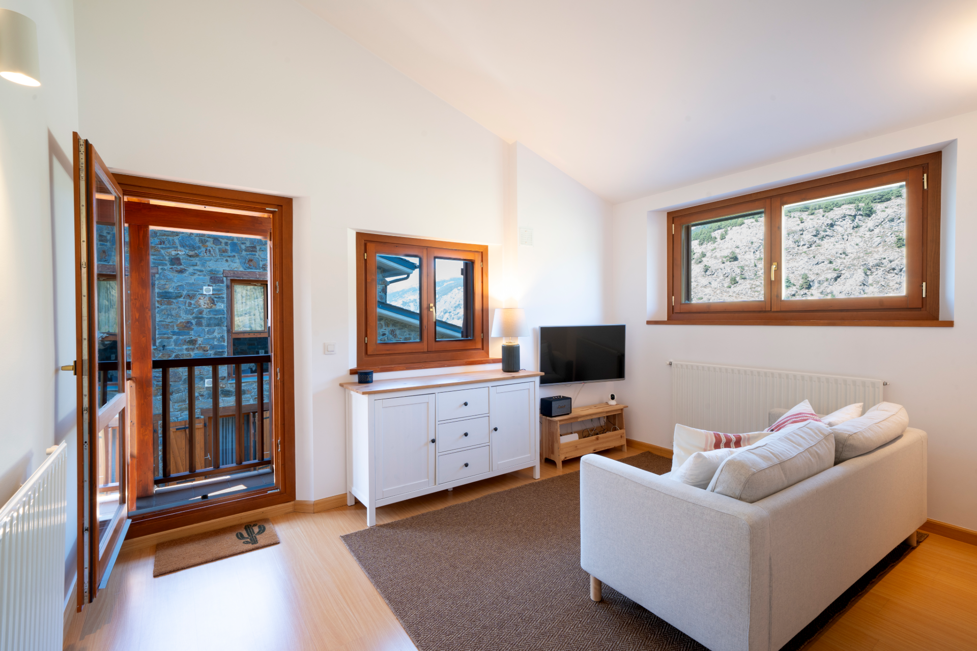 Bright living room with a white sofa, wooden sideboard, TV, and an open door leading to a balcony with mountain views. [AI-generated text]