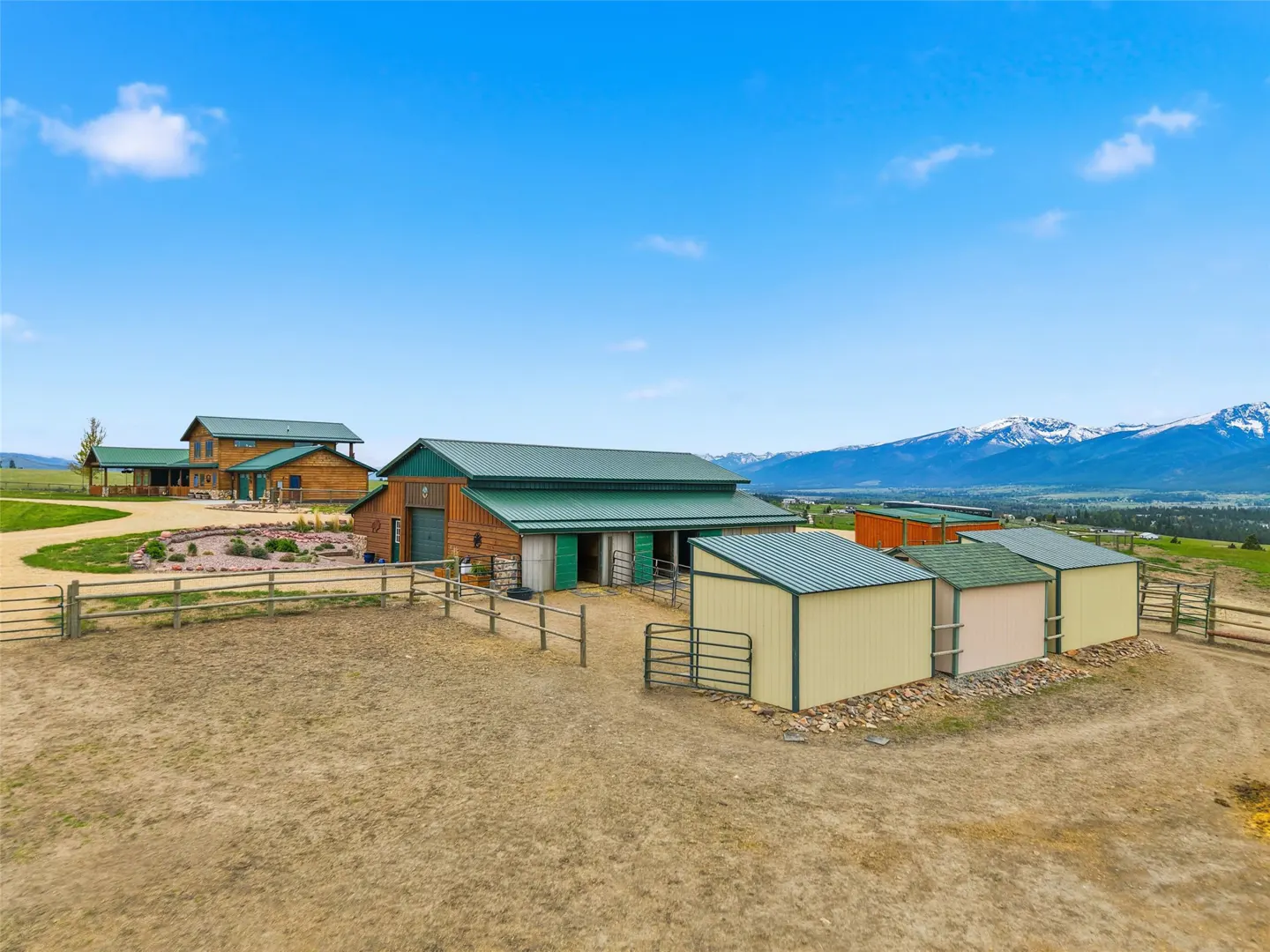 A ranch with a barn, sheds, and a house with a green roof, set against a backdrop of mountains and a blue sky.