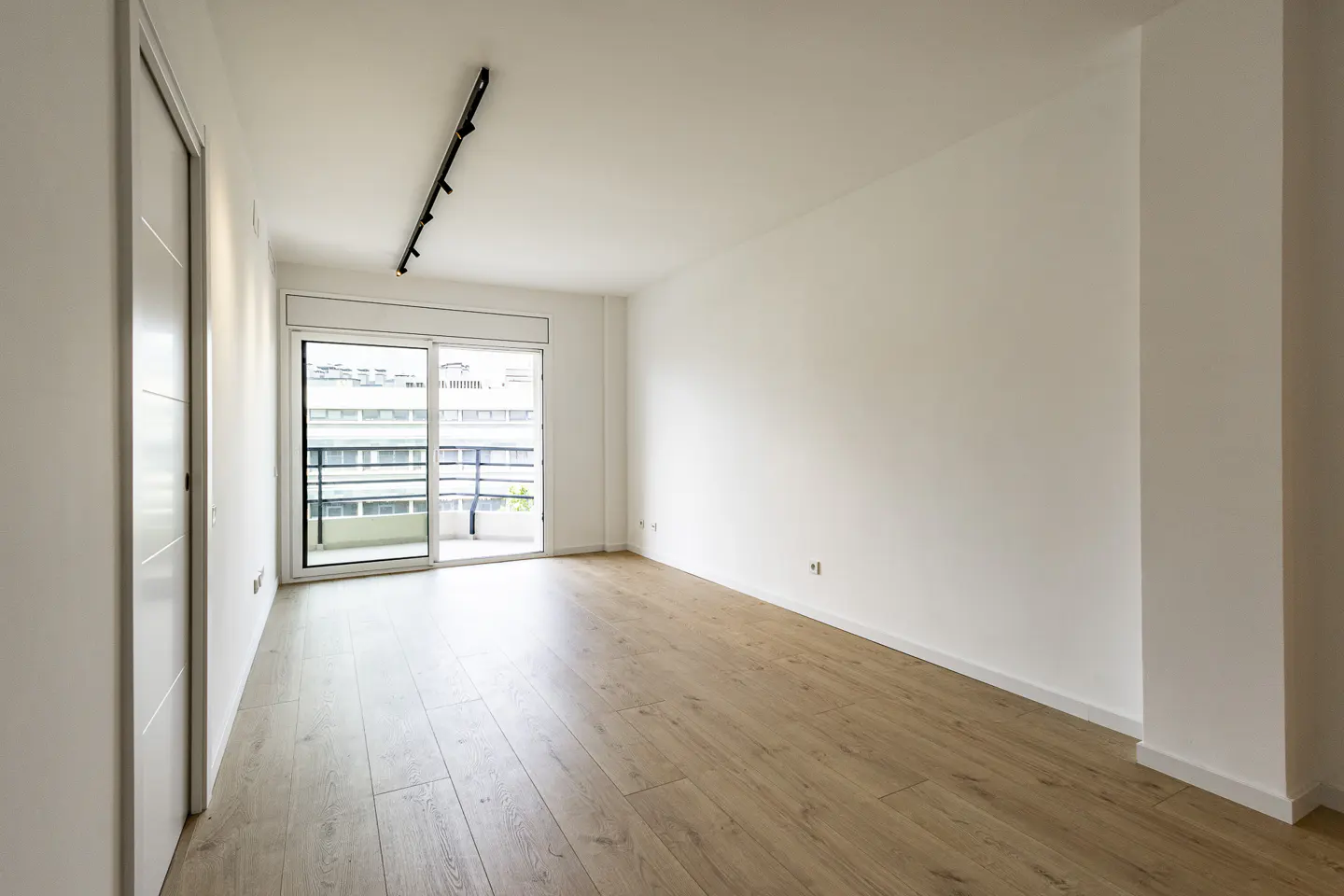 Bright, empty room with light wood floors, white walls, and a sliding glass door to a balcony. A black track light is on the ceiling.