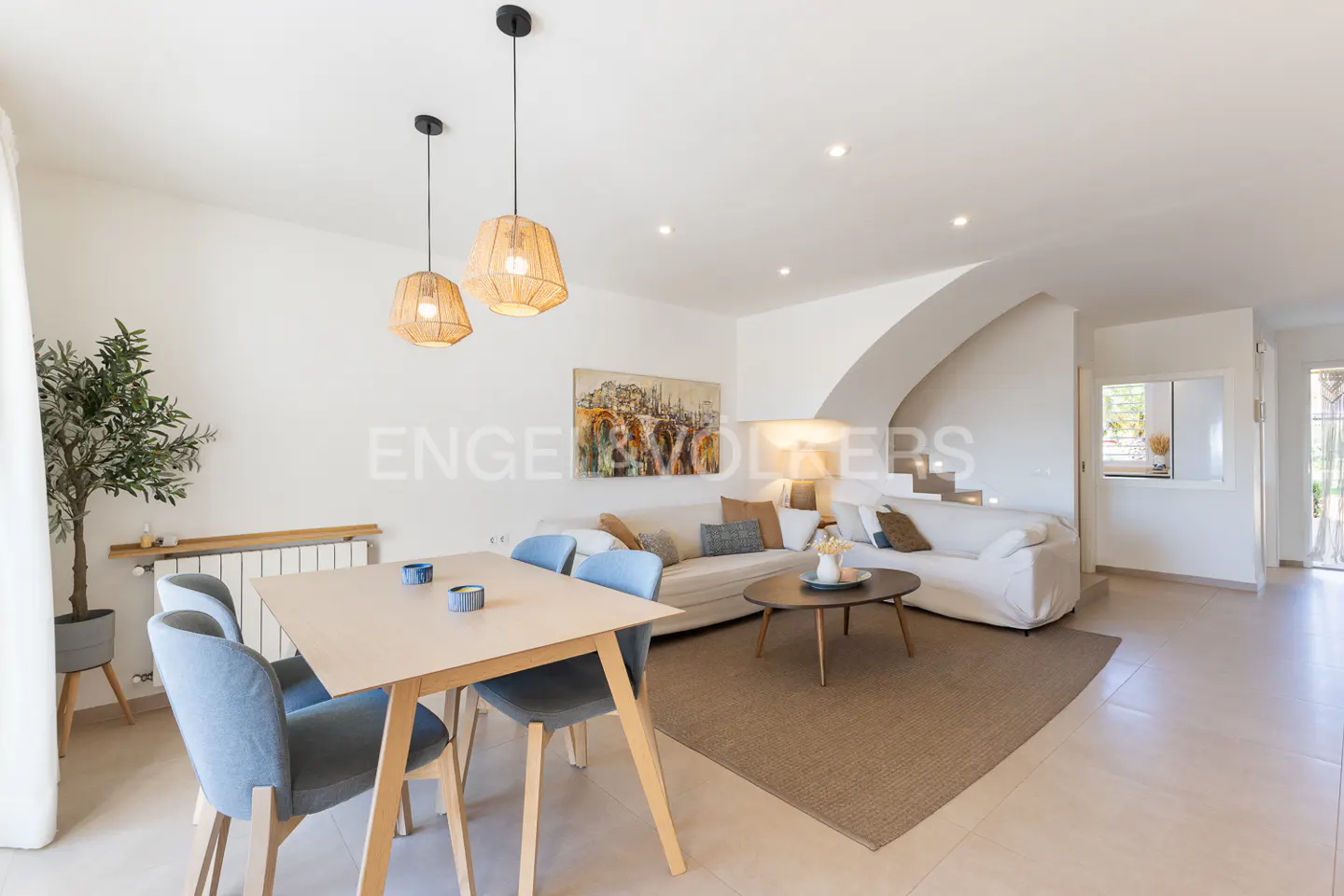 Bright, open-plan living space with dining table, blue chairs, white sofa, and woven pendant lights. Neutral tones and modern design.