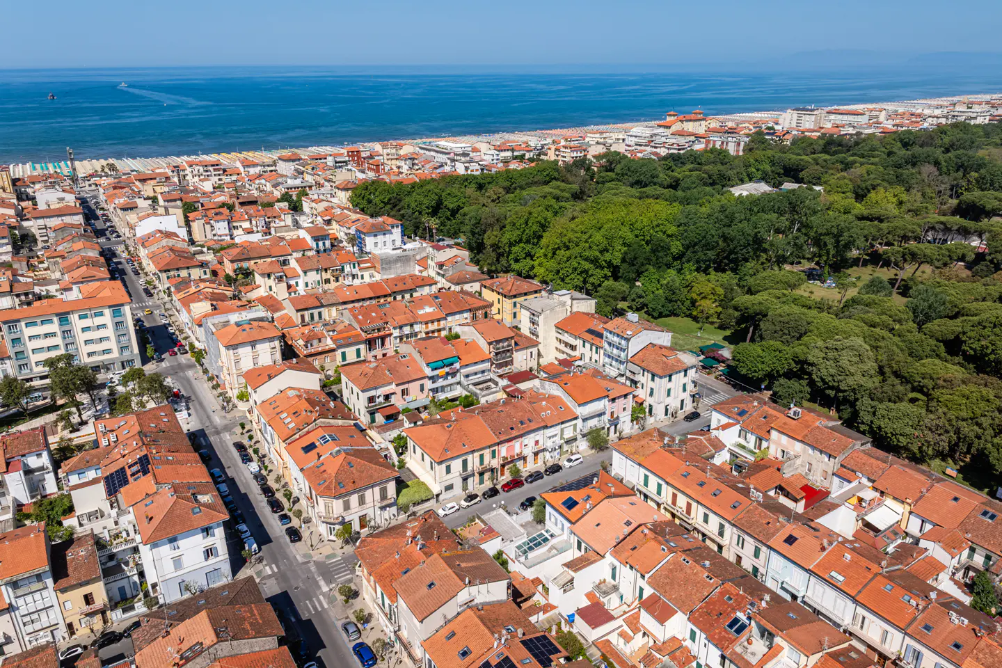 Aerial view of a coastal town with red-tiled roofs, green trees, and the blue sea in the background.