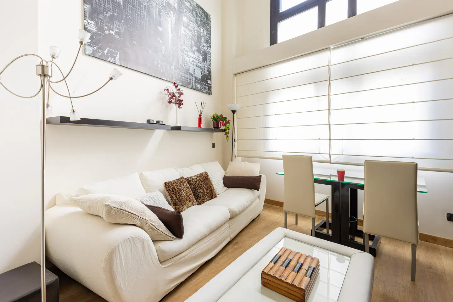 Bright living room with a white sofa, leopard print pillows, and a glass coffee table with a wooden box. A black and white city print hangs above.