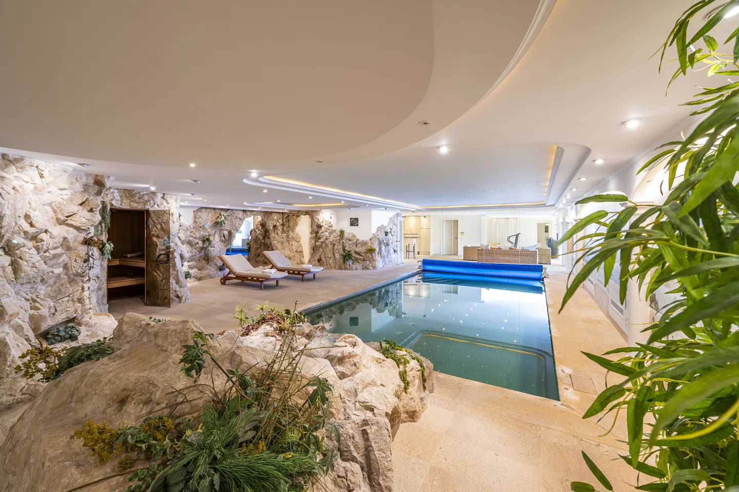 Indoor pool area with stone walls, sauna, and lounge chairs. The pool has a blue cover, and plants add a natural touch.