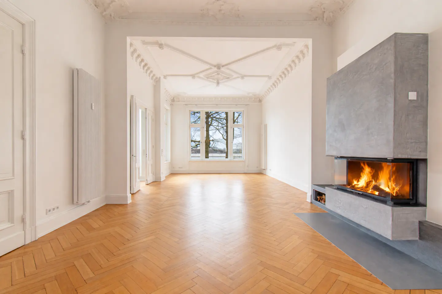Bright, empty room with herringbone wood floors, white walls, and a modern gray fireplace with a fire burning inside.