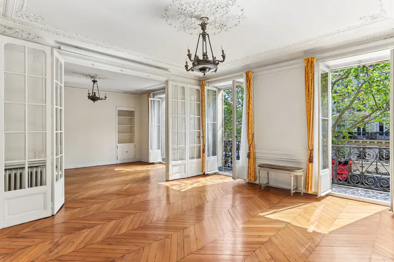 Bright, empty room with herringbone wood floors, white walls, and open French doors to a balcony with trees. A chandelier hangs from the ornate ceiling.