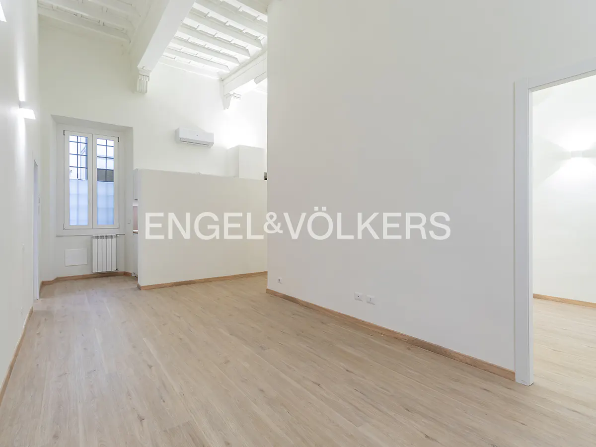 Bright, empty room with light wood floors, white walls, and white ceiling beams. A window is on the left, and a doorway is on the right. Engel & Völkers logo is in the center.