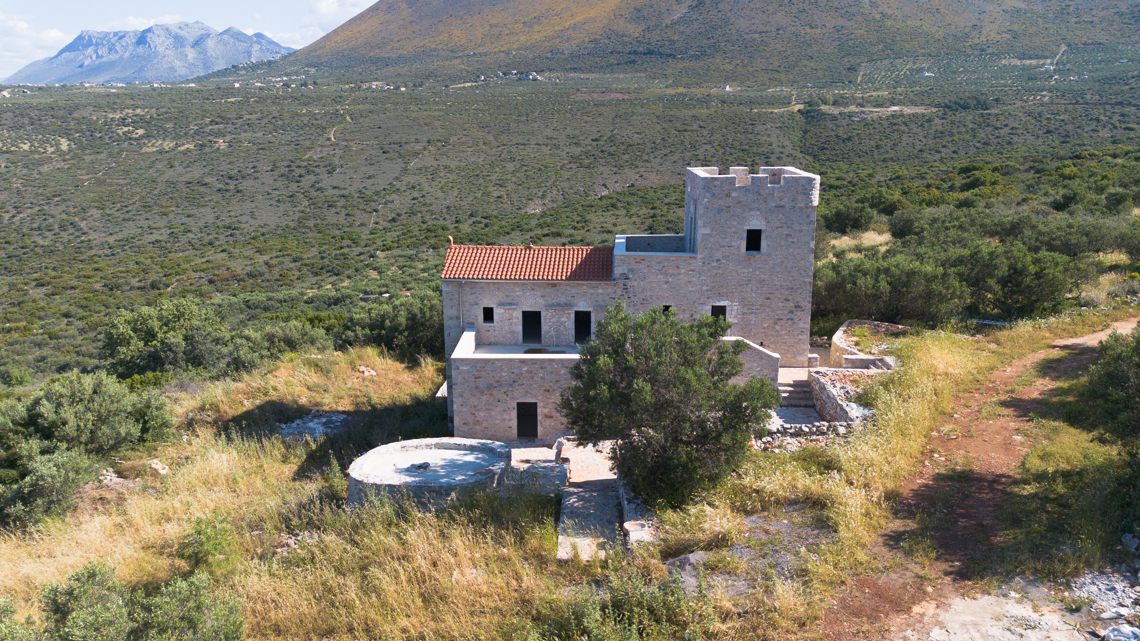 Stone house with red tile roof and tower, surrounded by green trees and mountains in the background. [AI-generated text]
