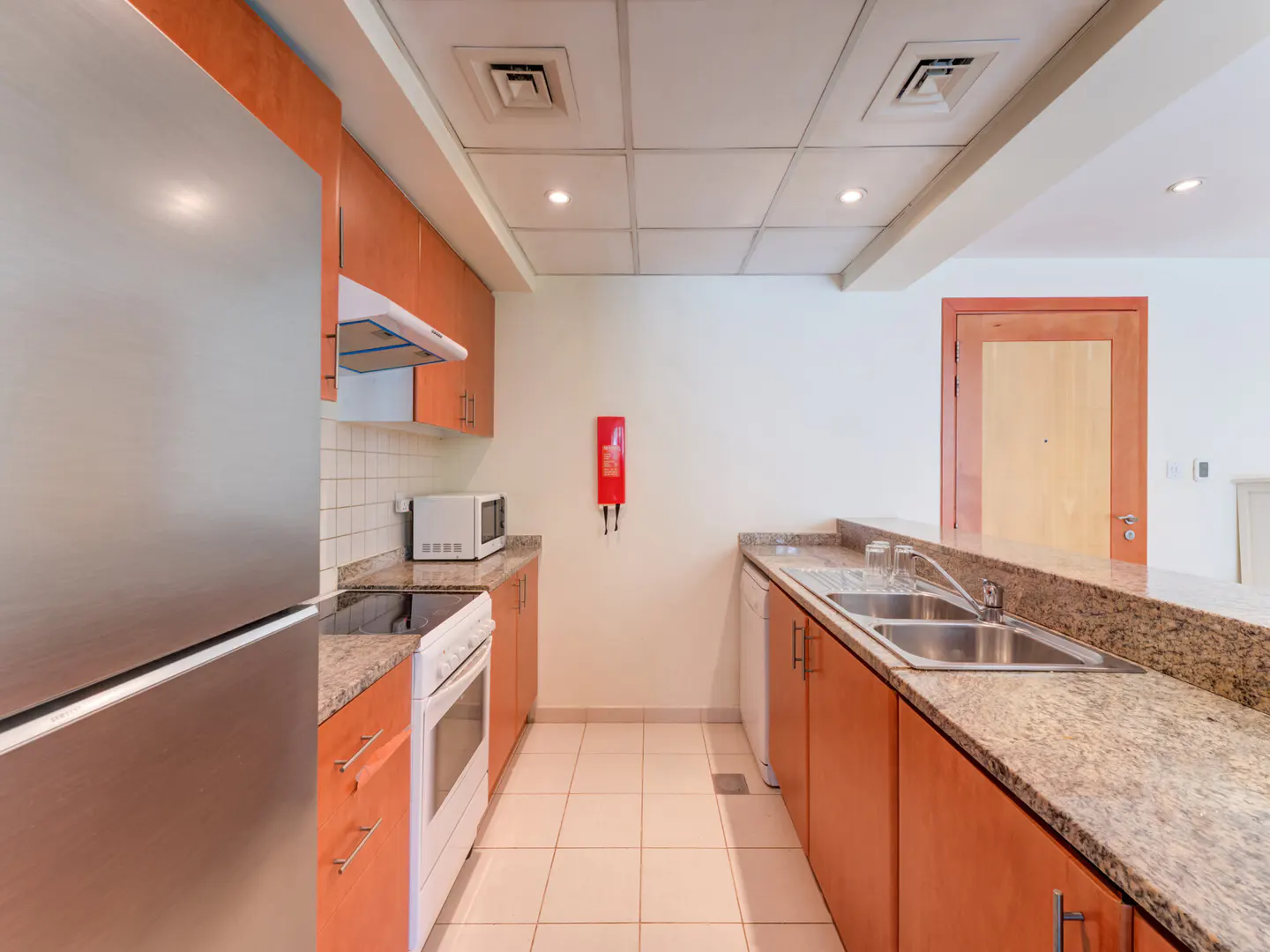 A kitchen with orange cabinets, granite countertops, and stainless steel appliances. A red fire blanket hangs on the wall.