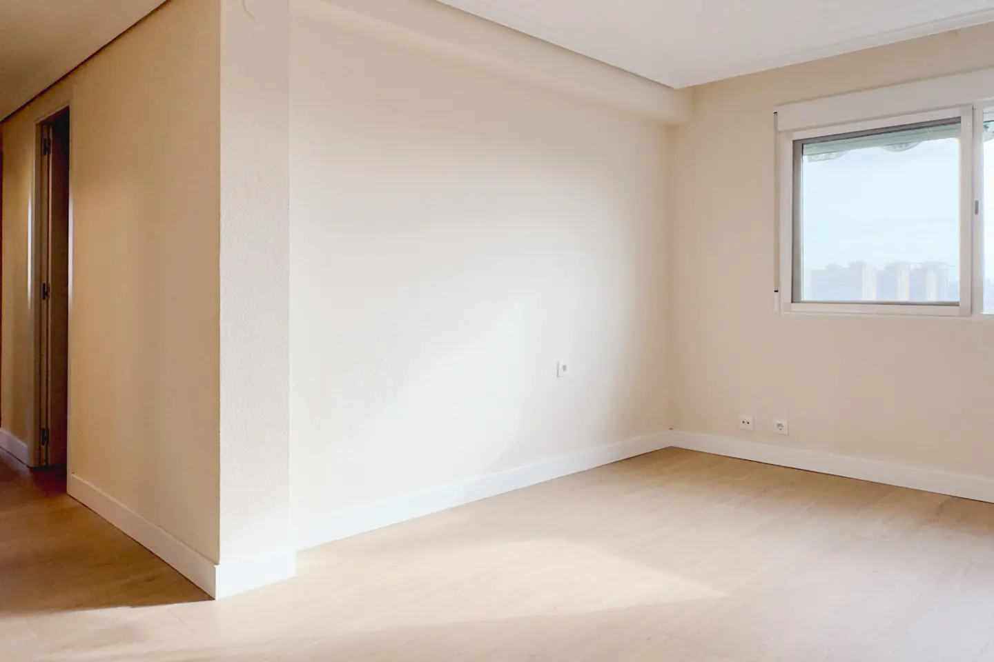Empty room with light wood floors, beige walls, and a window showing a city skyline.