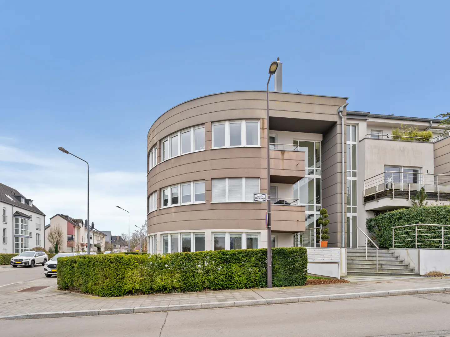 Exterior view of a modern, multi-story apartment building with a curved facade, balconies, and manicured hedges.