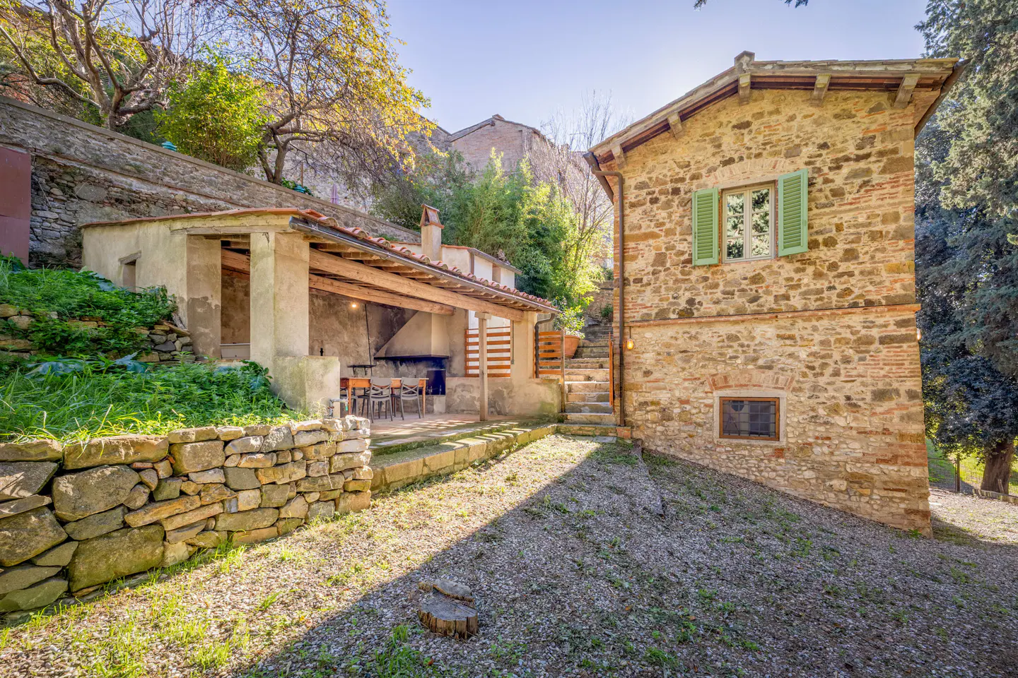 Rustic stone house with green shutters, a covered patio, and a stone retaining wall in a lush, sunlit garden setting.