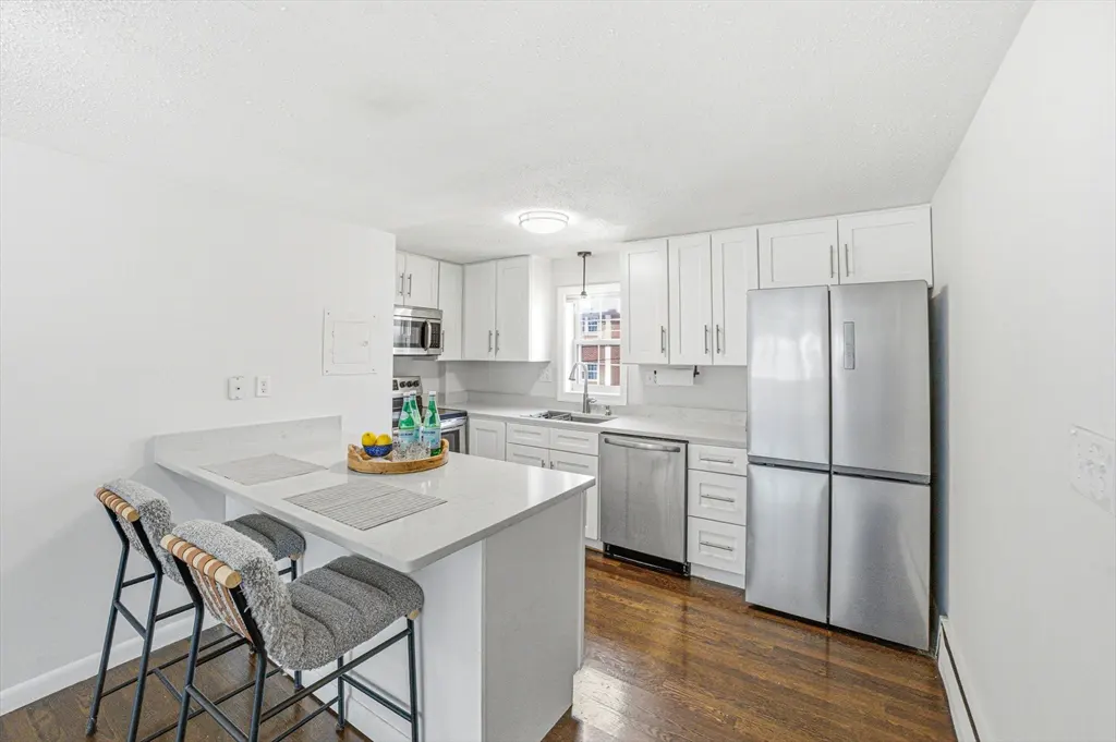 Bright kitchen with white cabinets, stainless steel appliances, and dark wood floors. A breakfast bar with two stools faces the kitchen.