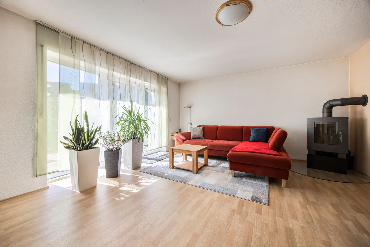 Bright living room with wood floors, a red sectional sofa, a wood-burning stove, and potted plants by a large window.