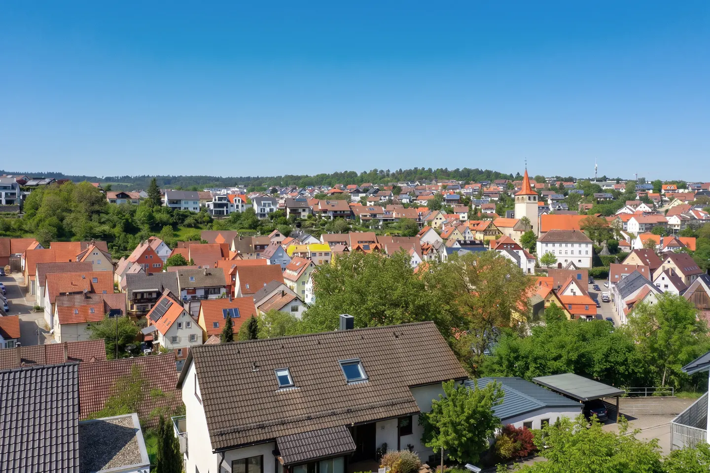 A sunny aerial view of a European town with red-tiled roofs, green trees, and a church with an orange spire.