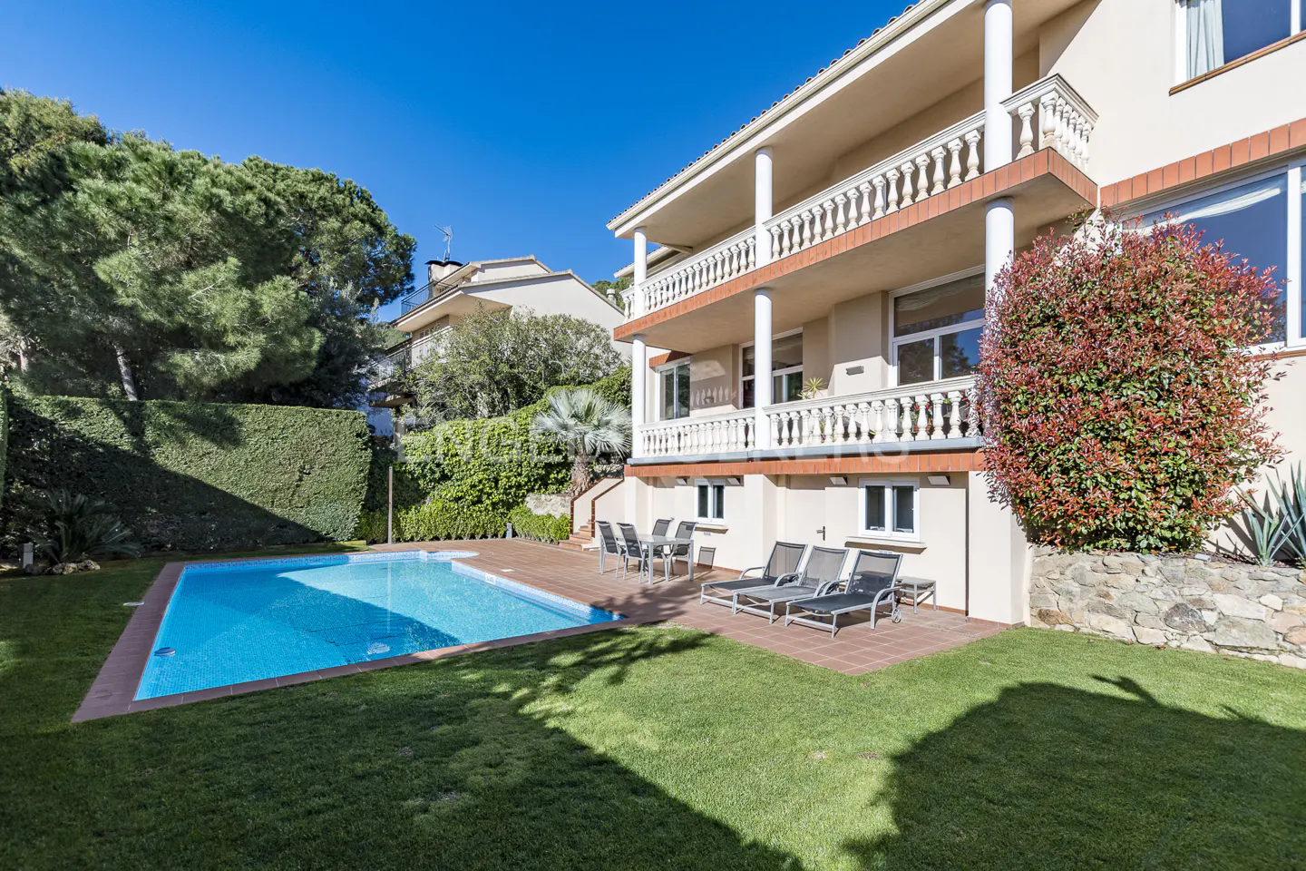 Backyard view of a two-story beige house with a blue pool, lounge chairs, and a balcony with white columns.