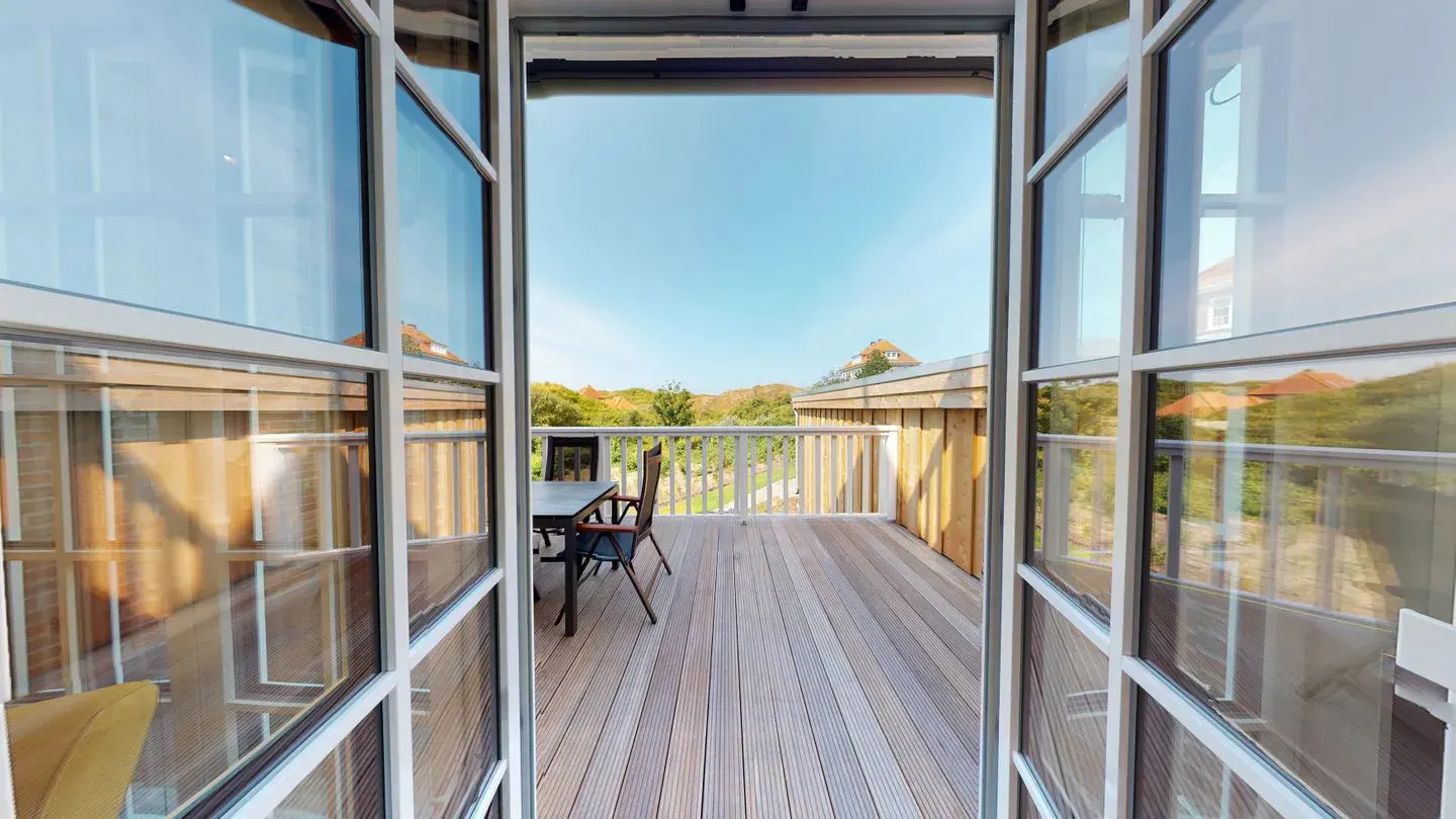 View from open white French doors onto a wood deck with table and chairs, white railing, and green trees under a blue sky.
