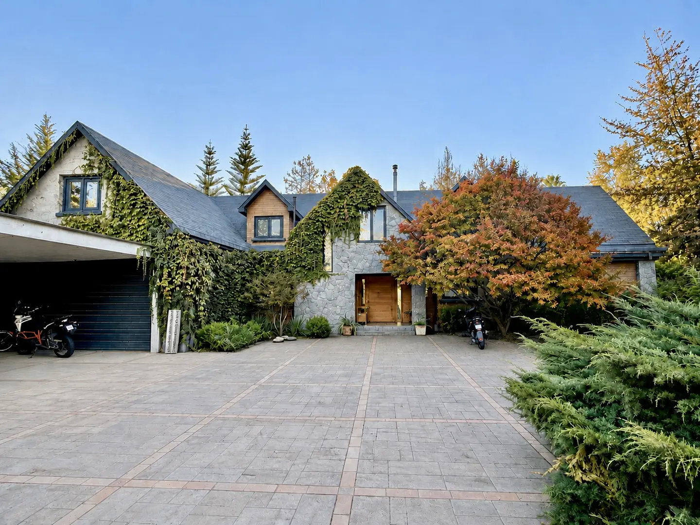 A two-story house with a gray stone facade, a dark roof, and ivy growing on the walls. A motorcycle is parked in the driveway.