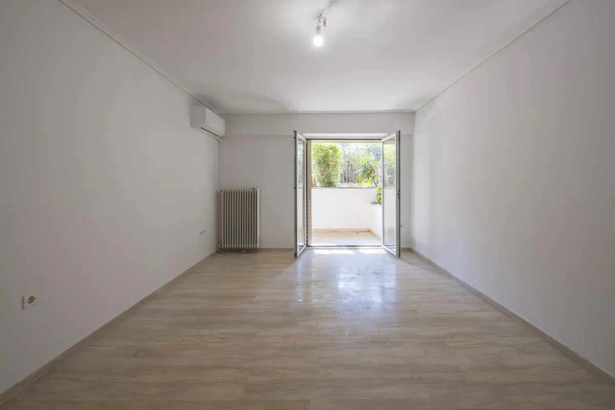 Bright, empty room with white walls, light wood floors, and open glass doors to a balcony with greenery. Radiator on the left wall.