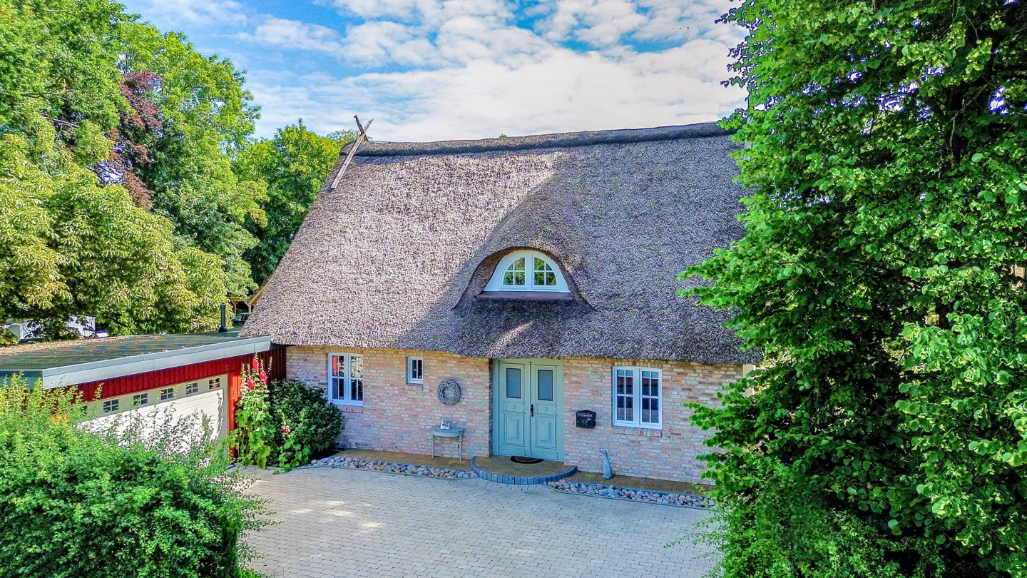 Exterior view of a tan brick house with a thatched roof, light blue door, and green trees.