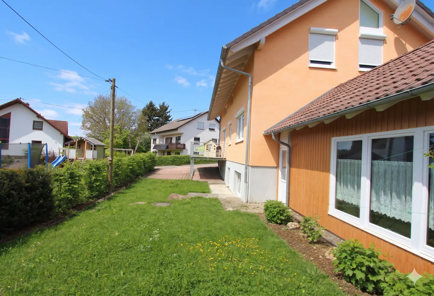A view of a house with an orange exterior and a brown roof, surrounded by a green lawn and trees under a blue sky.