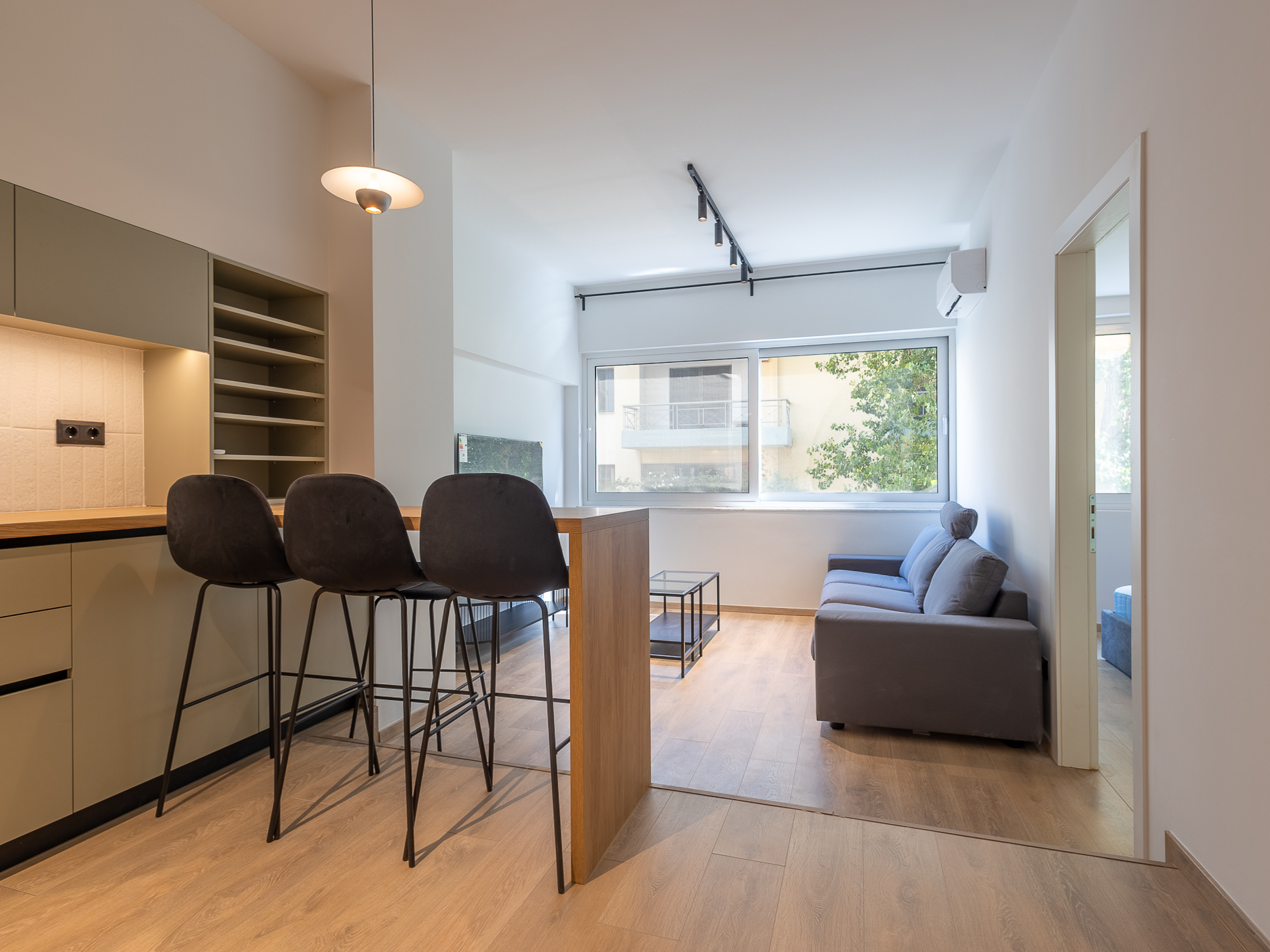 Modern open-plan apartment with a kitchen island, three bar stools, a grey sofa, and light wood flooring. [AI-generated text]