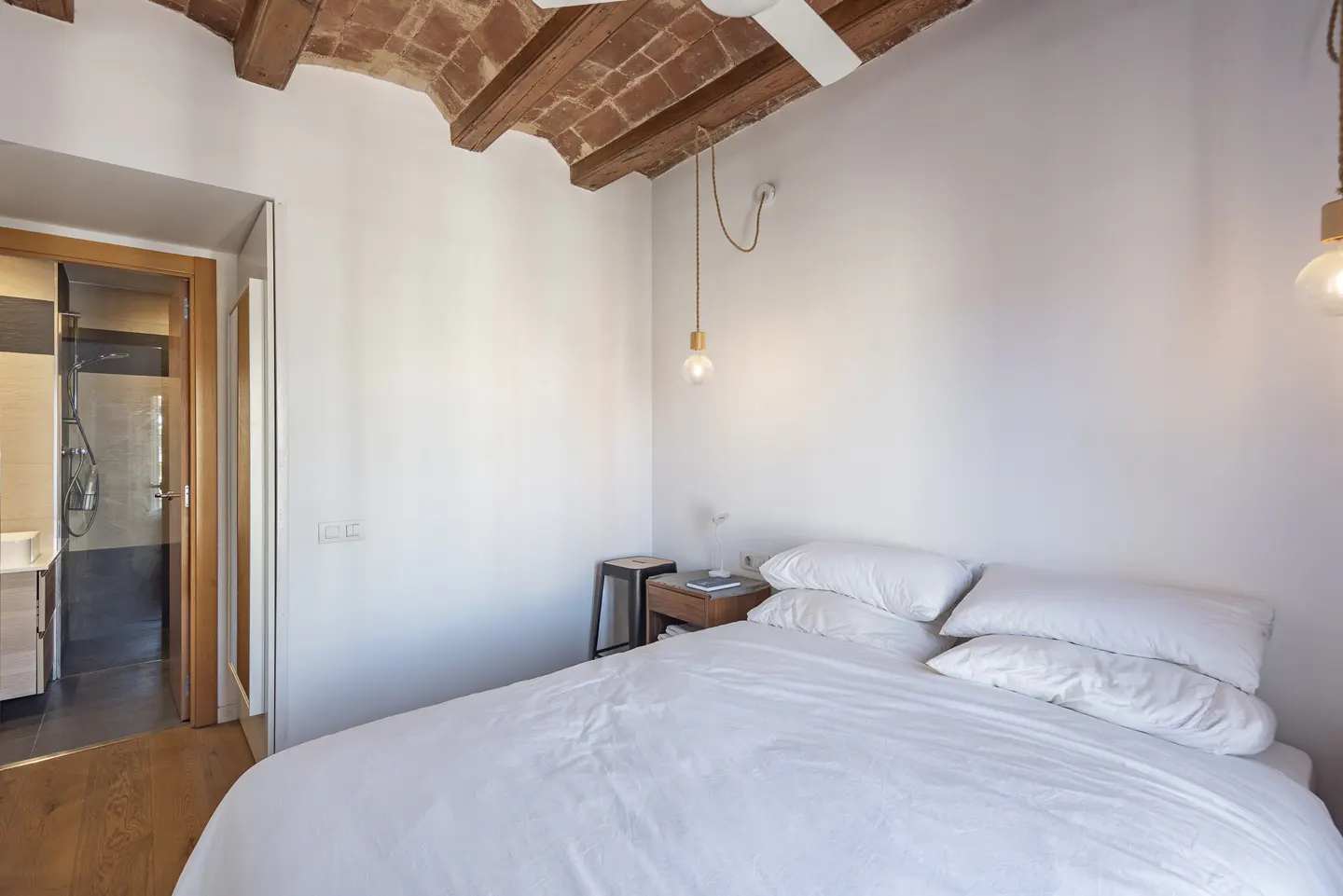 Bedroom with white walls, a bed with white linens, and a bathroom visible through an open door. The ceiling has exposed brick and wooden beams.