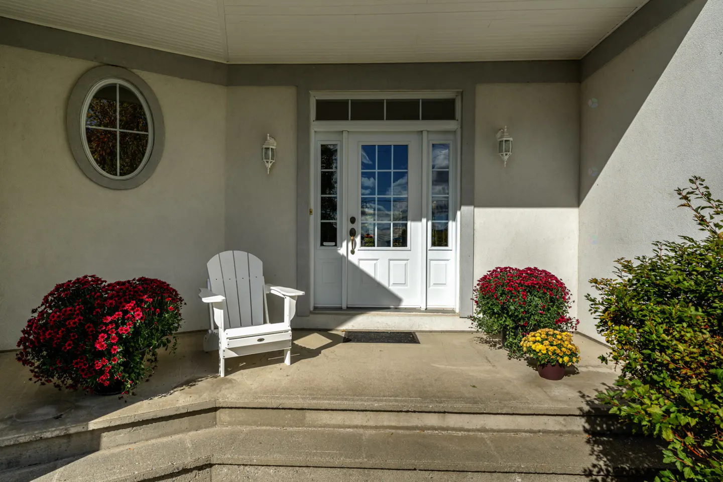 Front entrance of a white house with a white door, chair, red flowers, and a round window.