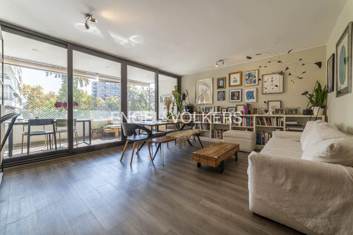 Bright living room with wood floors, a white sofa, a dining table, and a balcony with a view of trees and buildings.