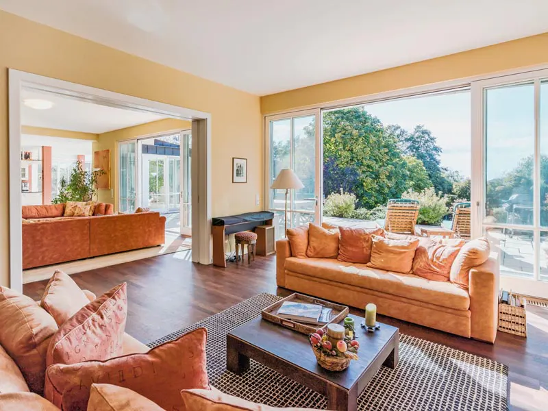 Living room with orange sofas, wood floors, and a large window overlooking a green yard. A piano sits near the window.
