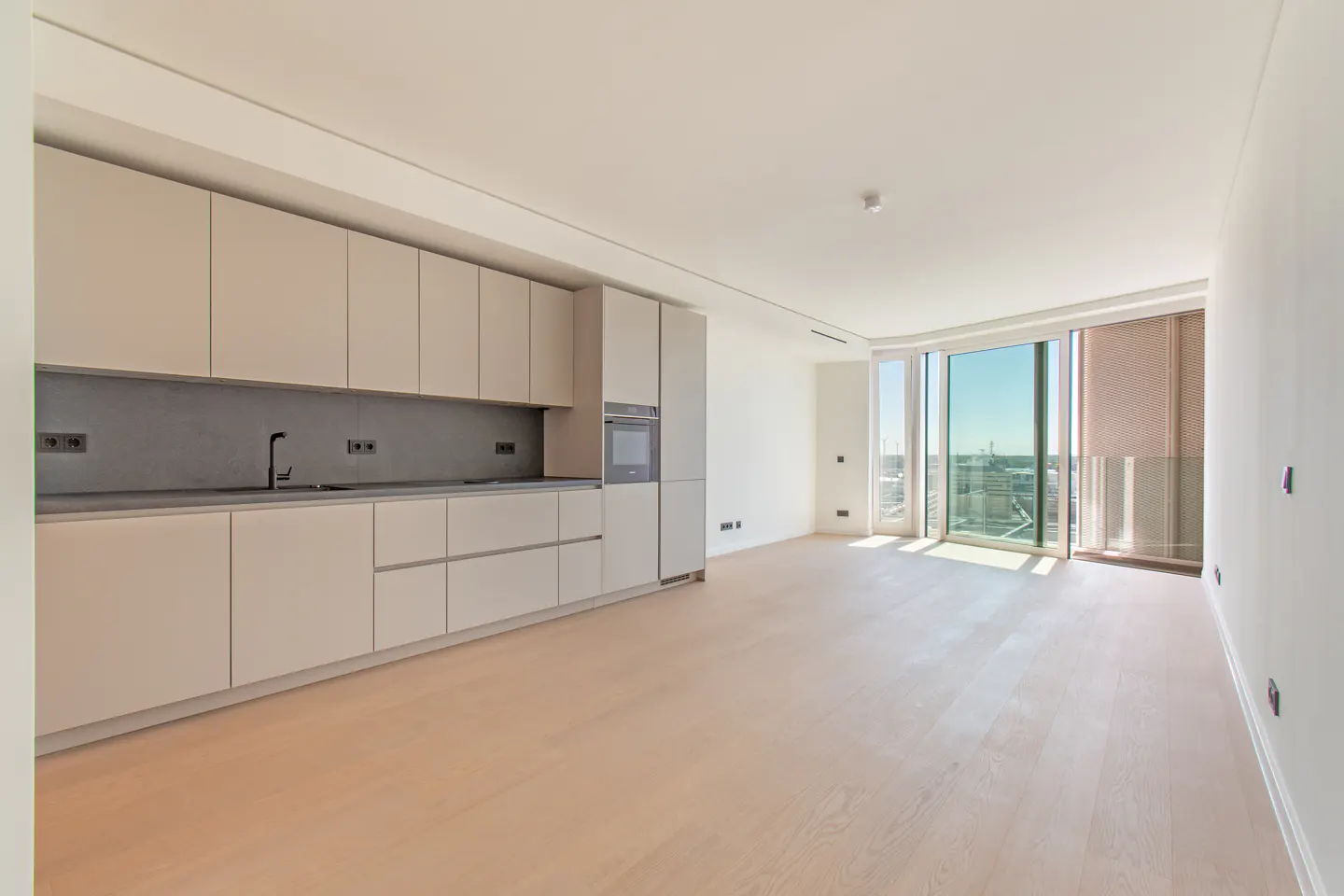 A modern, empty apartment with light wood floors, white walls, and a beige kitchen with a gray backsplash. Large windows offer a city view.