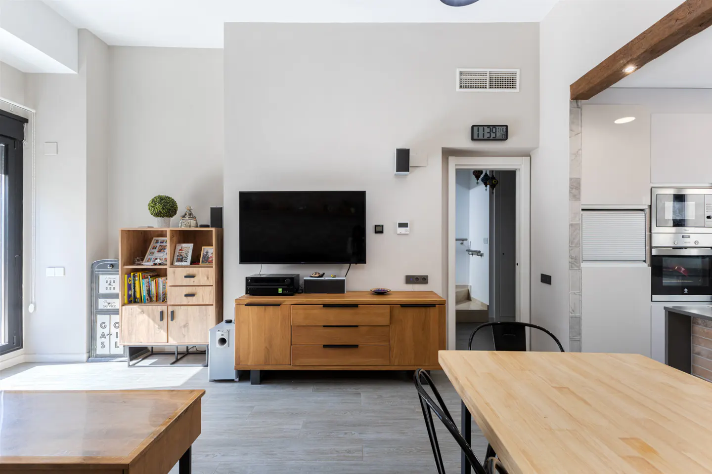 Bright, modern living room with a TV on a wooden console, a bookshelf, and a wooden table with black chairs.