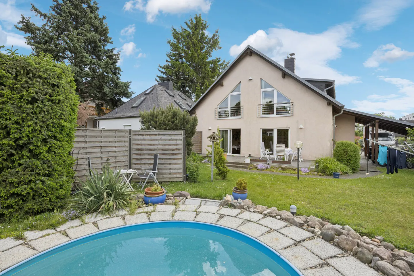 Backyard of a beige house with a round pool, stone patio, green lawn, and wooden privacy fence under a blue sky.