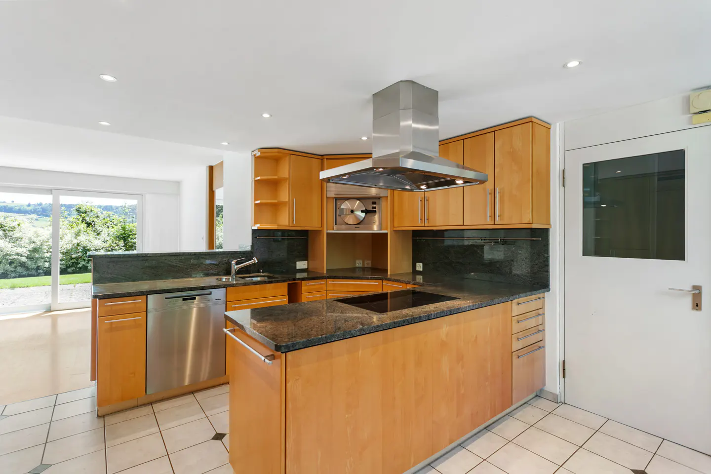 Modern kitchen with light wood cabinets, dark granite countertops, stainless steel appliances, and a view of the outdoors.