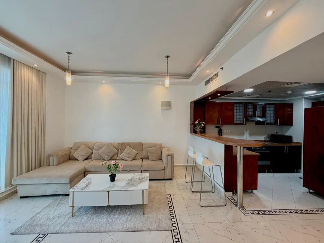 Modern living room with a beige sectional sofa, marble coffee table, and an open kitchen with dark wood cabinets and bar stools.