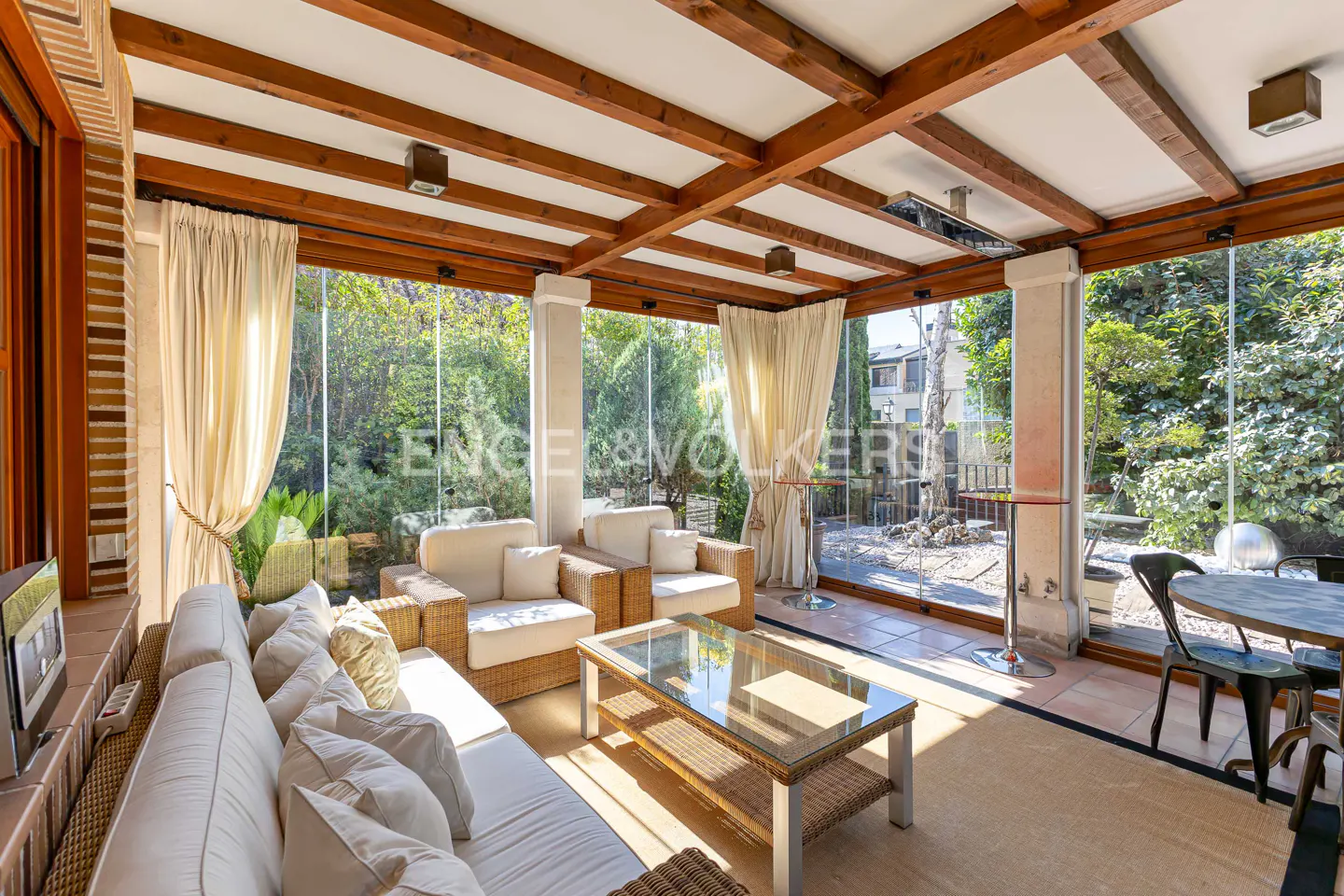 Bright sunroom with white sofas, wicker furniture, and glass walls overlooking a lush green garden. Wooden beams accent the ceiling.