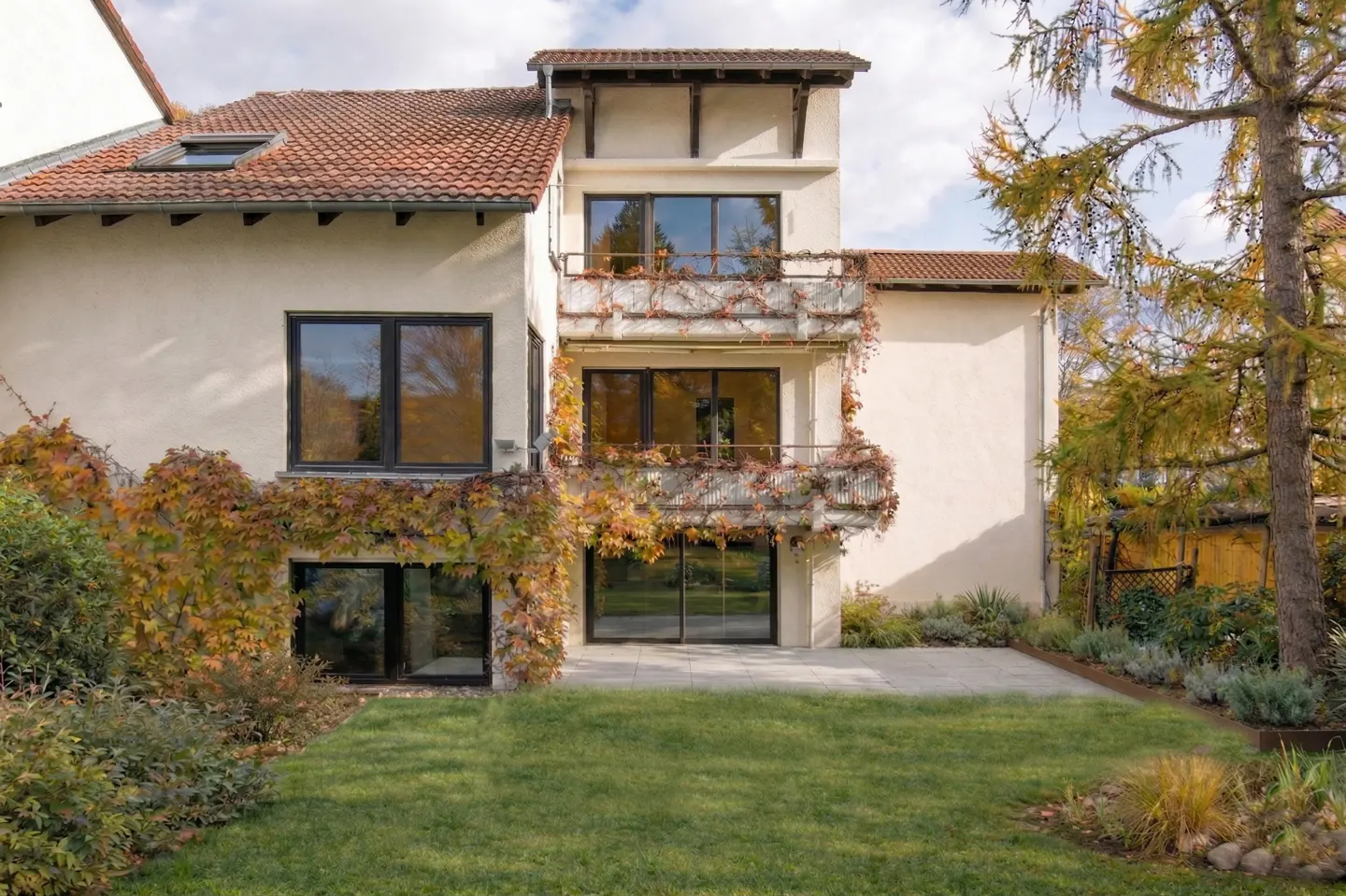Back view of a three-story house with balconies and a green lawn. Vines with orange leaves climb the building.
