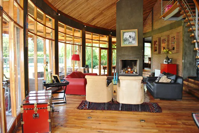 Open-concept living room with wood floors, a fireplace, and large windows. Two beige chairs face the fireplace. A red couch and black sofa are also visible.