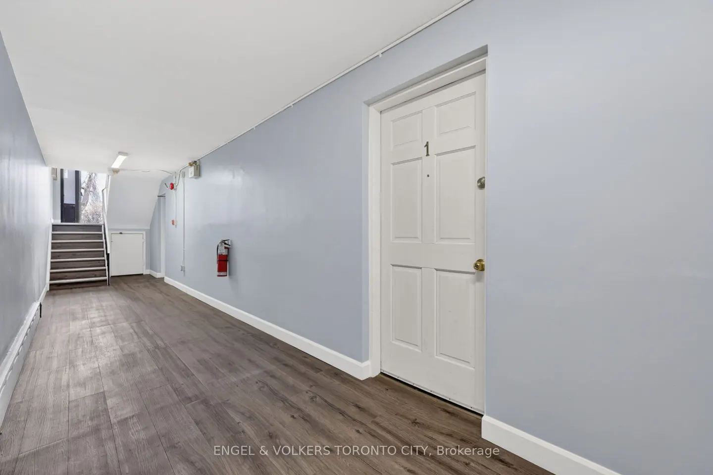 Hallway with light blue walls, wood floors, and a white door labeled "1". A red fire extinguisher is mounted on the wall. Stairs are visible in the background.