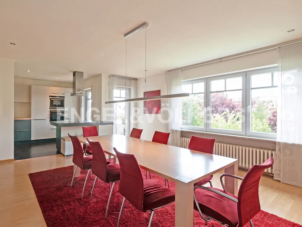 Dining room with a long wooden table, six red chairs on a red rug, and an open-plan kitchen in the background.