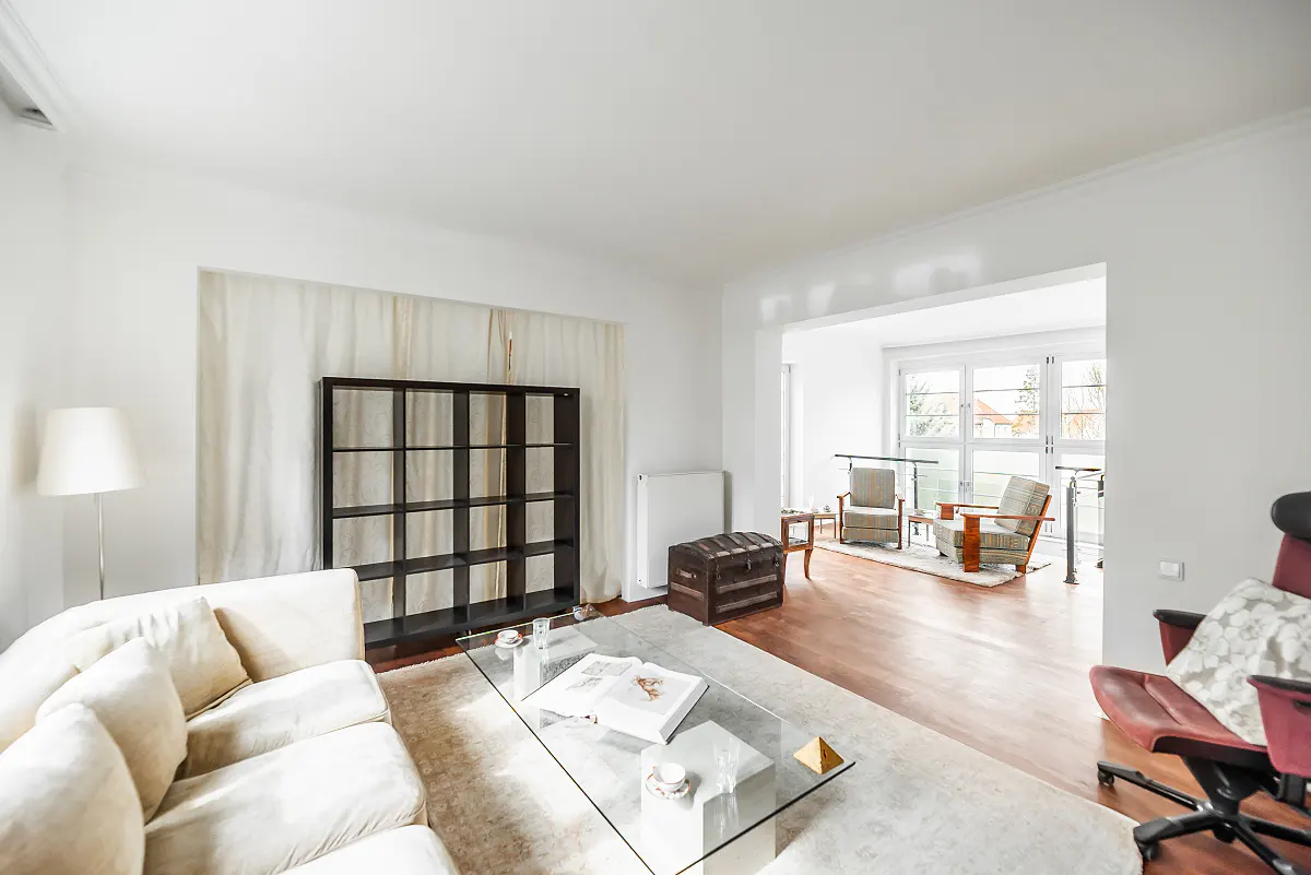 Bright living room with white walls, a cream sofa, a glass coffee table, and a black bookshelf. A doorway leads to another seating area.