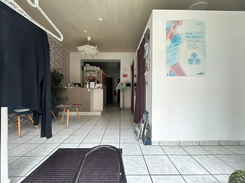 Interior view of a spa reception area with white tiled floors, a service desk, a waiting chair, and a promotional poster.