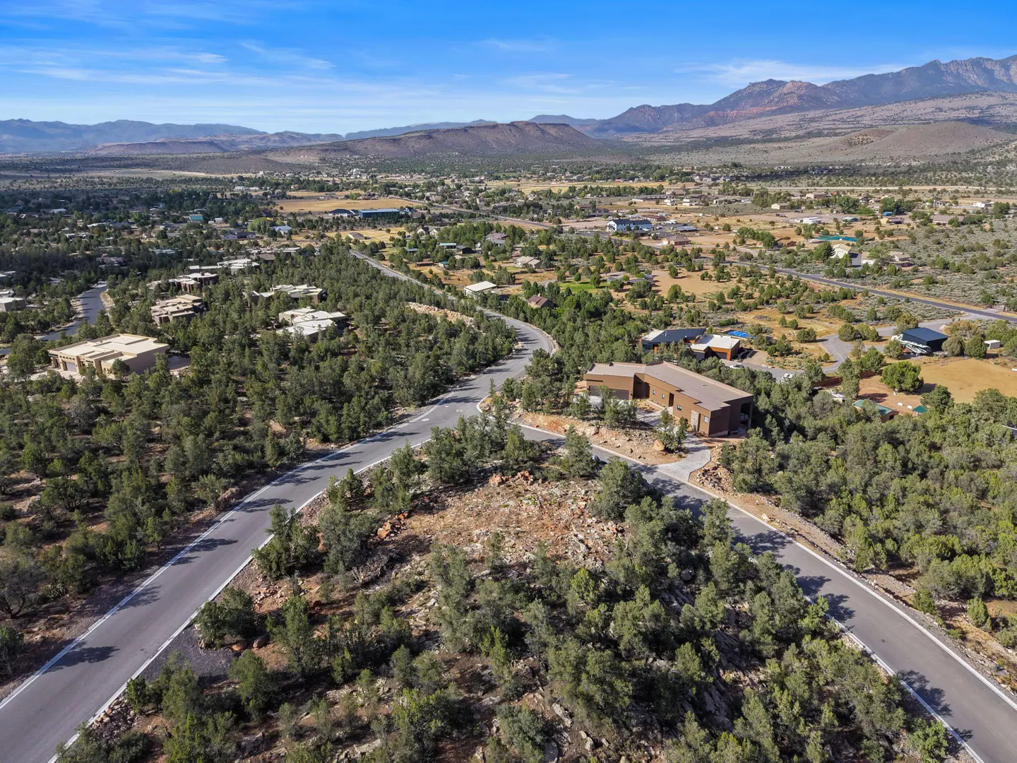 Aerial view of a brown house surrounded by green trees, a road, and mountains in the background under a blue sky.