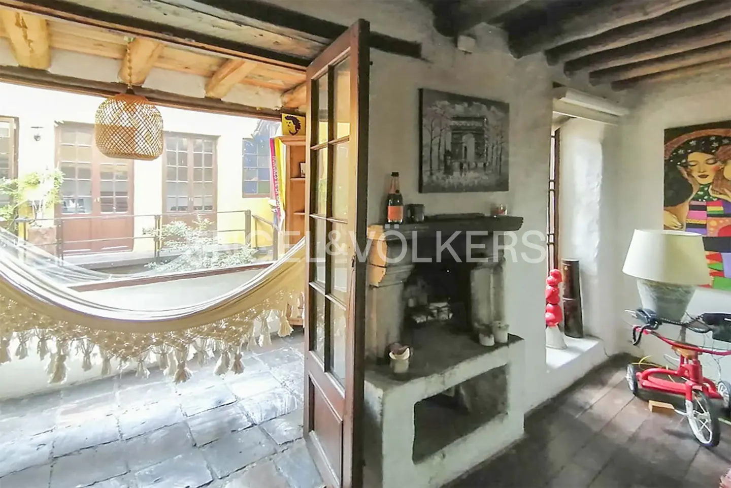 Interior view of a room with a stone fireplace, open doors to a courtyard with a hammock, and a child's tricycle.