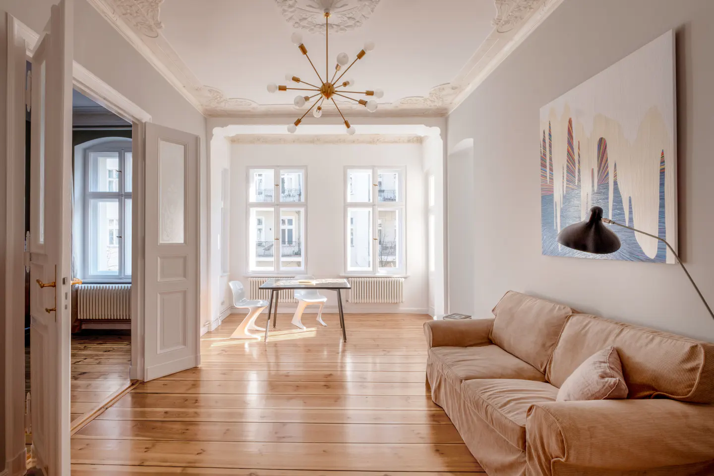 Bright living room with wood floors, beige sofa, and modern art. A table and chairs sit by the windows, with an open doorway to another room.