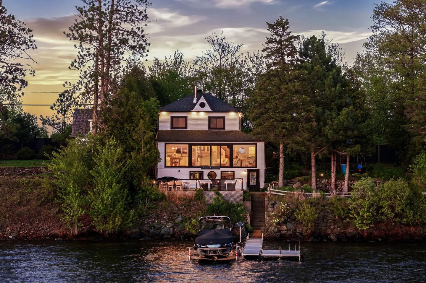Two-story white house with black trim on a lake. A boat is docked at the pier. Trees surround the house.