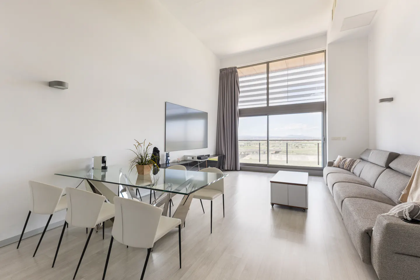 Bright living room with a glass table, white chairs, gray sofa, and large window with a view.
